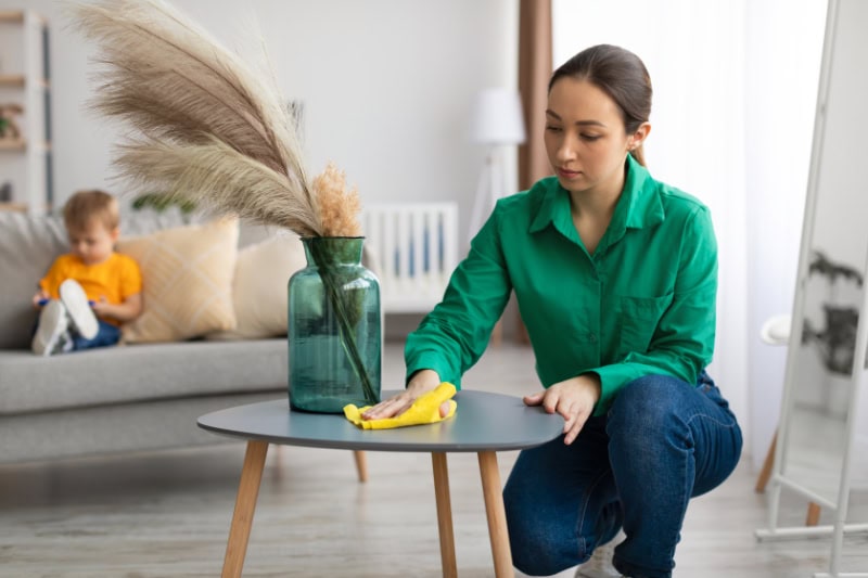 Duct Cleaning. Spring cleaning. Young mom dusting off coffee table and tidying up in living room while her toddler son watching cartoon on cellphone, sitting on sofa on background.